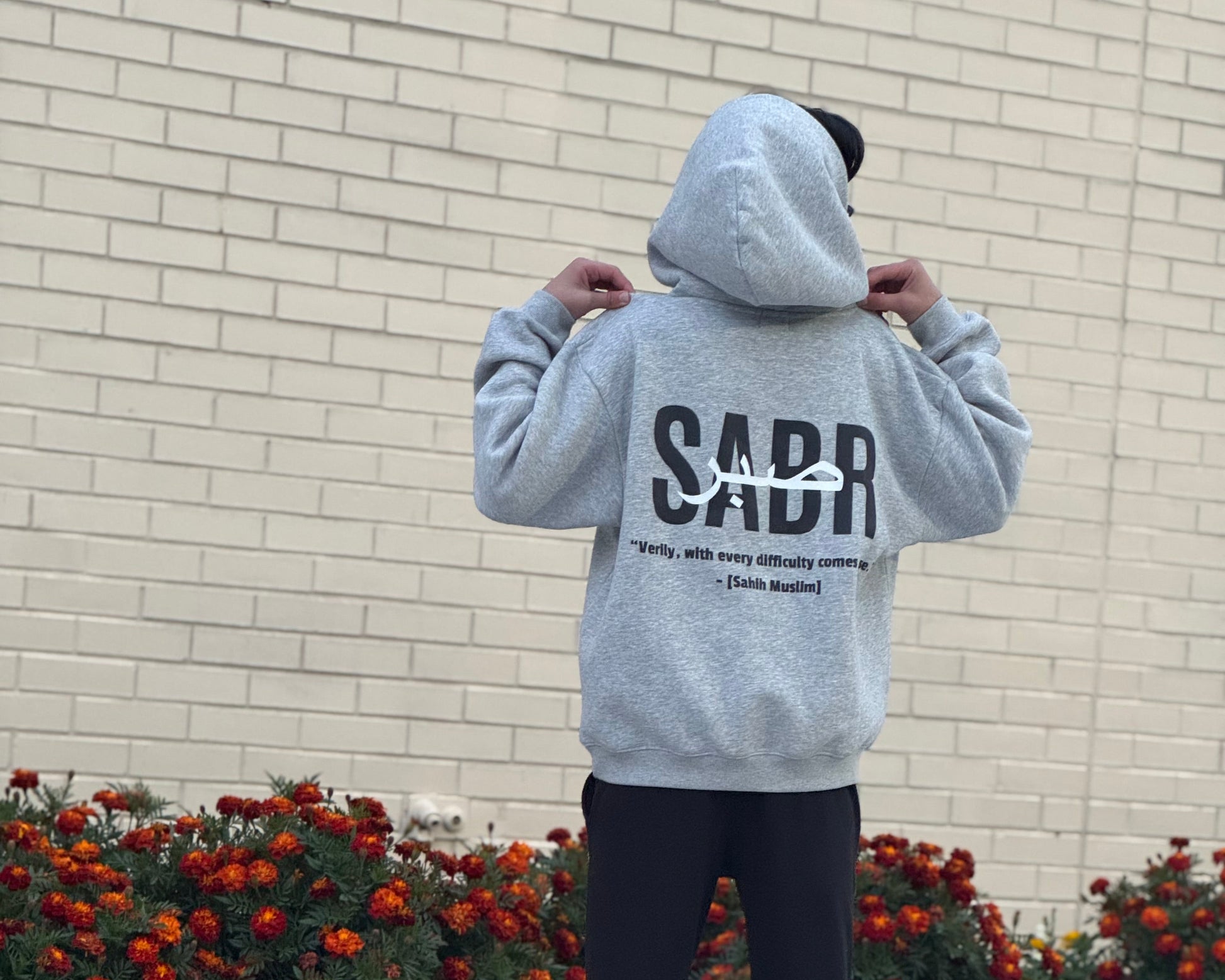 Person wearing a gray hoodie with 'SABR' printed on the back, standing in front of a brick wall with flowers.